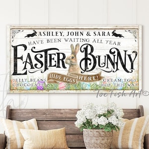 May include: A white wooden sign with the text "Ashley, John & Sara have been waiting all year Easter Bunny" featuring a cartoon bunny rabbit and colourful Easter eggs. The sign is hanging on a white wall above a wooden bench with cushions and a potted plant.