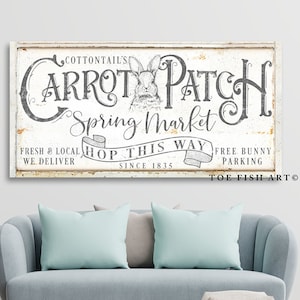 May include: A rectangular, distressed white sign with the text "Cottontail's Carrot Patch Spring Market" and a rabbit illustration. The sign also includes the phrases "Fresh & Local," "Hop This Way," and "Free Bunny Parking." The sign is mounted above a blue sofa.