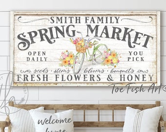 May include: A white wooden sign with black text that reads "Smith Family Spring Market". The sign features a bicycle with a basket of flowers and the text "Open Daily" and "You Pick". The sign also includes the text "Seeds, Stems, Blooms, Bouquets" and "Fresh Flowers & Honey". The sign is made by Toe Fish Art.