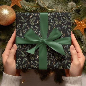May include: A wrapped gift with a black background and a botanical pattern of green and gold leaves. The gift is tied with a dark green satin ribbon and bow. The gift is surrounded by Christmas decorations.