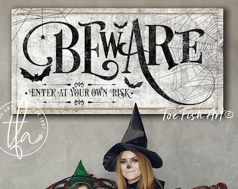 May include: A black and white sign with the text "Beware Enter at your own risk" with a spider web and bats. Two people dressed as witches are sitting on a white couch with pumpkins in their hands. The sign is from Toe Fish Art.