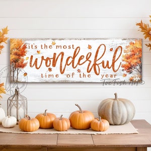 May include: A white wooden sign with the text "it's the most wonderful time of the year" in orange lettering. The sign is decorated with fall leaves and a small pumpkin. The sign is hanging on a white wood wall.