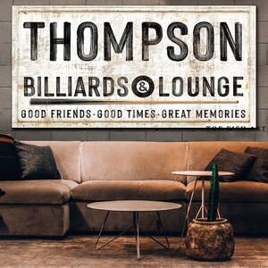 May include: A white wooden sign with black lettering that reads "Thompson Billiards & Lounge" with the text "Good Friends. Good Times. Great Memories" below. The sign has a distressed look and a brown pool cue stick is resting on the sign.