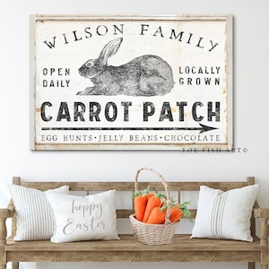 May include: A white sign with black text that reads "WILSON FAMILY", "OPEN DAILY", "LOCALLY GROWN", "CARROT PATCH", "EGG HUNTS JELLY BEANS CHOCOLATE", and "TOE FISH ART". A black and white illustration of a rabbit is above the text. The sign is hanging on a white wall above a wooden bench with two cushions and a basket of carrots.