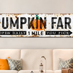 May include: A white, rusty metal sign with black text that reads "Pumpkin Farm" with a small orange pumpkin graphic. The sign also says "Open Daily 1 Mile You Pick" with a black arrow pointing to the right. The sign is on a white wall.