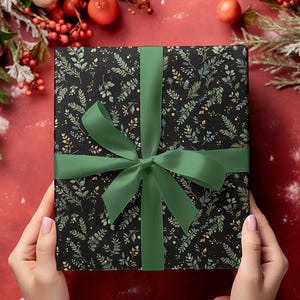 May include: A wrapped gift box with a black background and a botanical pattern of green and gold leaves. The box is tied with a green satin ribbon and bow. The gift is held by a person.
