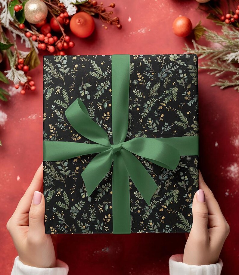 May include: A wrapped gift box with a black background and a botanical pattern of green and gold leaves. The box is tied with a green satin ribbon and bow. The gift is held by a person.