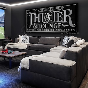 May include: A black and white sign with the text "Welcome to the Theater & Lounge" in a decorative font. The sign also includes the text "Movies, Popcorn, Drinks, Games" and the signature "loe Fish Art".