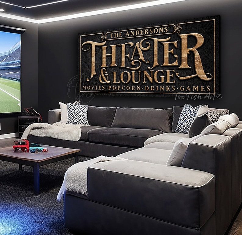 May include: A black and gold sign with the text "The Andersons' Theater & Lounge" and the words "Movies, Popcorn, Drinks, Games" below. The sign is hanging on a black wall in a home theater room.