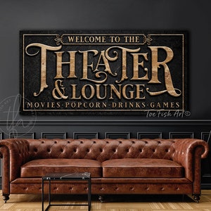 May include: A black and gold sign with the text "Welcome to the Theater & Lounge" and "Movies Popcorn Drinks Games". The sign has a distressed wood texture and is hanging on a dark gray wall above a brown leather sofa.