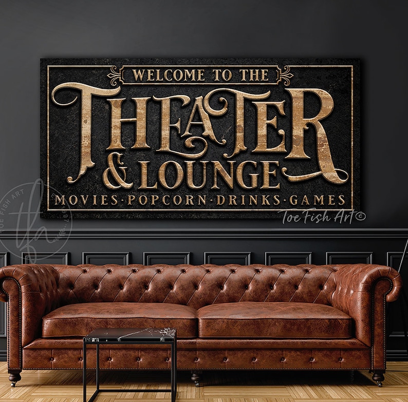 May include: A black and gold sign with the text "Welcome to the Theater & Lounge" and "Movies Popcorn Drinks Games". The sign has a distressed wood texture and is hanging on a dark gray wall above a brown leather sofa.