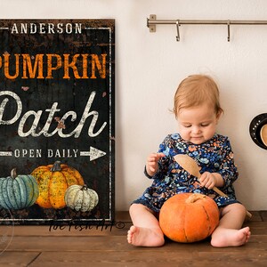 Op de afbeelding: Een zwart-wit bord met de tekst "Anderson Pumpkin Patch Open Daily" en een afbeelding van drie pompoenen. Een jong kind zit op een houten vloer met een pompoen voor zich.