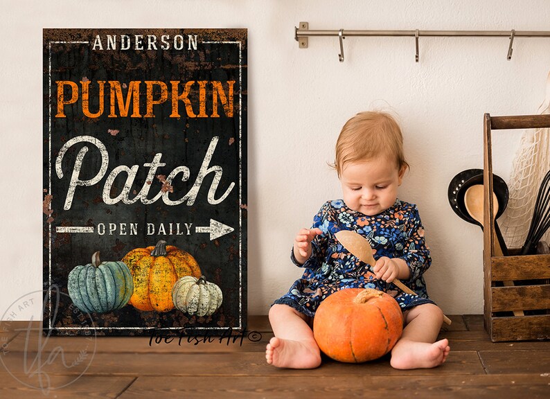 Op de afbeelding: Een zwart-wit bord met de tekst "Anderson Pumpkin Patch Open Daily" en een afbeelding van drie pompoenen. Een jong kind zit op een houten vloer met een pompoen voor zich.