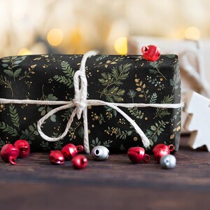 May include: A wrapped gift box with black wrapping paper featuring a green and gold botanical pattern. The box is tied with white twine and surrounded by red and silver bells on a dark wooden surface. A white wooden tree is in the background.