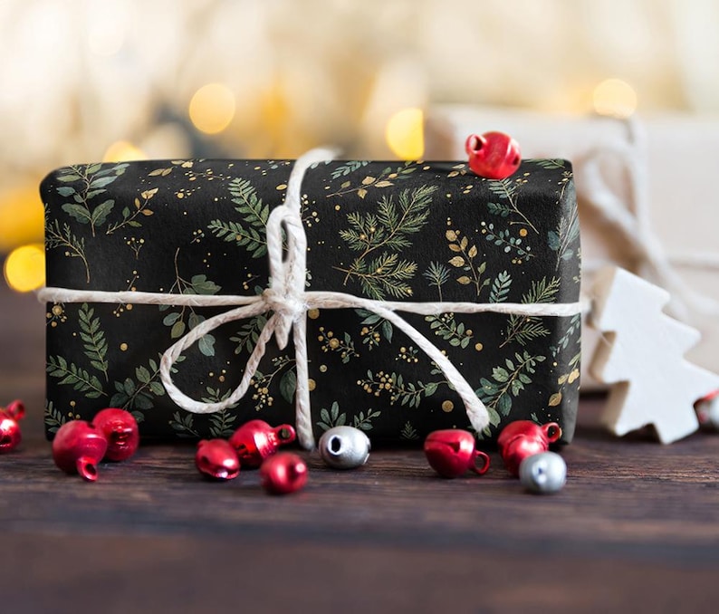 May include: A wrapped gift box with black wrapping paper featuring a green and gold botanical pattern. The box is tied with white twine and surrounded by red and silver bells on a dark wooden surface. A white wooden tree is in the background.