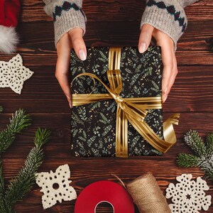 May include: A wrapped gift with a black floral pattern and gold ribbon is held by a person. The gift is surrounded by Christmas decorations, including ornaments, snowflakes, and evergreen branches. The scene is set on a wooden surface.