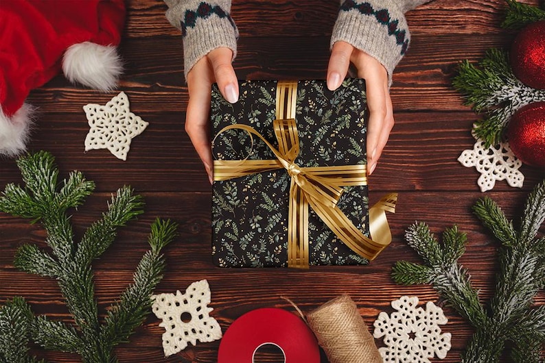May include: A wrapped gift with a black floral pattern and gold ribbon is held by a person. The gift is surrounded by Christmas decorations, including ornaments, snowflakes, and evergreen branches. The scene is set on a wooden surface.