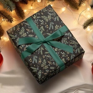 May include: A wrapped gift box with a dark background and a pattern of green and gold leaves. The box is tied with a wide, emerald green satin ribbon and bow. Christmas ornaments and evergreen branches are in the background.