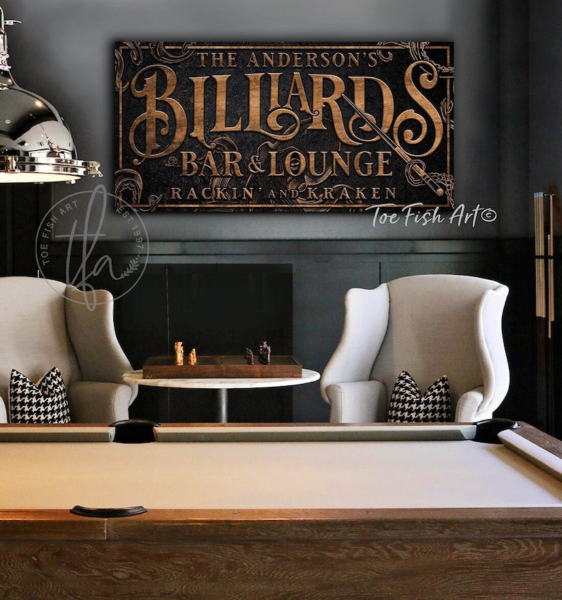May include: A wooden sign with gold lettering that reads "The Anderson's Billiards Bar & Lounge Rackin' and Kraken" with a vintage style design. The sign is hanging on a dark wall above a pool table.