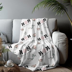Op de afbeelding: Een witte fleece deken met een bloemenpatroon en de tekst "Mom" die zich herhaalt. De tekst is zwart en de bloemen zijn roze en groen.