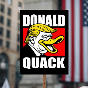 May include: A protest sign with the words "DONALD QUACK" in white capital letters. The sign features a cartoon illustration of a duck with yellow hair and a black suit, resembling a former U.S. president, set against a red background.