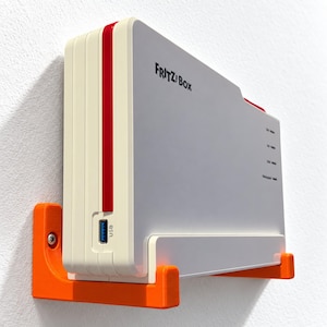 Puede incluir: Un router FRITZ!Box blanco y rojo montado en la pared mediante un soporte de plástico de color naranja brillante. El router cuenta con un puerto USB frontal, un panel lateral con el logotipo FRITZ!Box y cuatro pequeñas luces indicadoras.