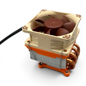 May include: A computer CPU cooler featuring a square beige fan with dark brown blades, mounted atop a silver aluminium heatsink with copper heat pipes. The assembly includes bright orange plastic mounting brackets and a black power cable.