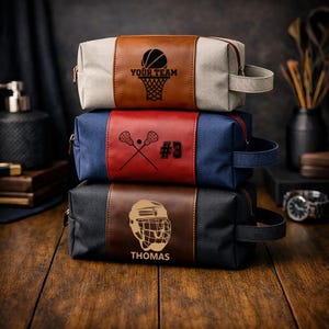 May include: Three canvas and leather toiletry bags stacked on a wooden surface. The top bag has a basketball and the text "YOUR TEAM". The middle bag has lacrosse sticks and the number "#3". The bottom bag has a hockey helmet and the name "THOMAS".