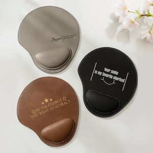 Op de afbeelding: Drie ergonomische muismatten in grijs, bruin en zwart. De grijze pad heeft de naam "Jasper" en de tekst "DON'T TOUCH MY KEYBOARD". De bruine pad heeft de tekst "BE THE ENERGY YOU WANT TO ATTRACT". De zwarte pad heeft de tekst "Your smile is my favorite shortcut".