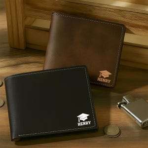 May include: Two leather wallets, one brown and one black, with white stitching. Each wallet features a graduation cap graphic and the name "HENRY". A silver lighter and coins are also visible.