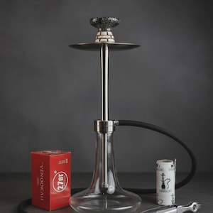 May include: A modern hookah set with a clear glass base, a silver metal stem, and a black hose. The set includes a red box with text, a small container, and metal tongs. The hookah has a dark, patterned top.