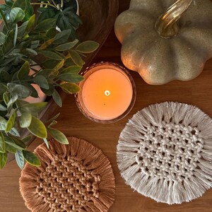 May include: Two round macrame coasters with fringed edges, one in a warm terracotta orange and the other in an off-white cream color, featuring a central woven knot pattern made from cotton cord, displayed on a wooden surface.