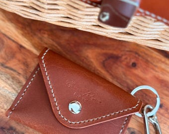May include: A brown leather key holder with white stitching and a silver snap closure. The key holder has a silver keyring and a clasp. The background includes a wooden surface and a woven leather bag.