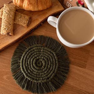 May include: A round, olive green macrame coaster featuring a tightly coiled center and a fringed outer edge. The coaster is crafted from cord, displaying a spiral pattern that radiates from the middle to the textured, knotted fringe border.