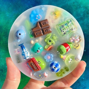 May include: A clear, circular resin piece containing various miniature charms. Features a blue cloud, chocolate bar, green bunny, and sweets. The background is a starry, blue and green gradient. The piece is held in a hand.
