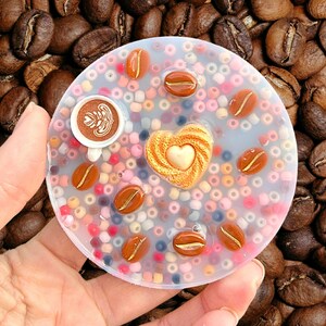 May include: A clear, round coaster decorated with coffee beans and a heart shape. The coaster includes a small coffee cup with latte art and is surrounded by colourful beads. The background is filled with coffee beans.