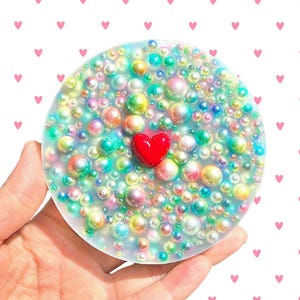 May include: A circular resin coaster featuring a translucent base embedded with numerous iridescent, multi-coloured spherical beads of varying sizes. A single, smooth, solid red heart-shaped charm is positioned in the centre of the coaster.