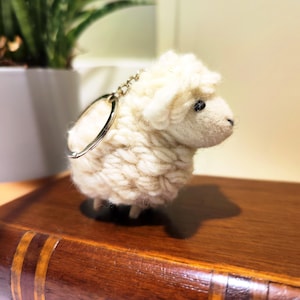 May include: A small, fluffy white sheep keyring with a silver ring. The sheep has a textured wool appearance and small black eyes. It is sitting on a wooden surface, with a plant in the background.