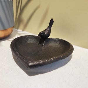 May include: A black, heart-shaped trinket dish with a small bird figurine on the edge. The dish has a textured rim and sits on a white surface. The bird is facing left, and the dish is made of metal.