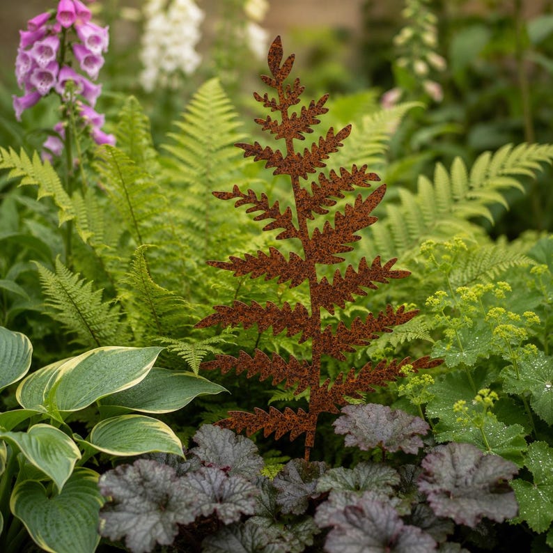 May include: A decorative metal garden stake shaped like a fern frond with a textured, rusted brown finish. The piece features a central stem with symmetrical, jagged-edged leaves extending outward, designed to stand upright among green foliage.