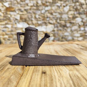 May include: A cast iron watering can doorstop. The doorstop is dark brown with a textured surface. The watering can has a handle and a spout. The doorstop is on a wooden surface.