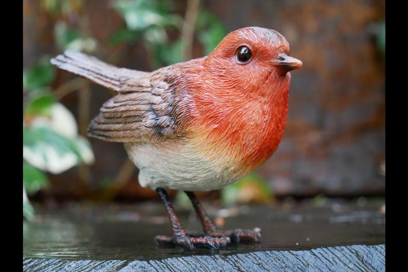 May include: A detailed figurine of a European robin. The bird has a red breast, brown wings, and a white belly. It is perched on a surface, with its legs visible. The robin's eyes are dark and it has a small beak.