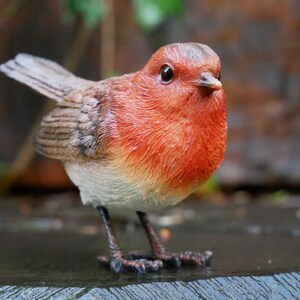 May include: A detailed resin figurine of a European robin. The bird has a bright orange breast, a grey-brown back, and a small, dark beak. Its wings are slightly spread, and it stands on two tiny legs. The figurine is likely made of resin or a similar material.