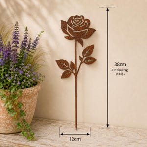 May include: A rust-colored metal rose garden stake featuring a cutout flower design with leaves on a thin stem. The total height is 38 cm including the stake, and the width is 12 cm. The metal has a textured, weathered brown finish throughout.