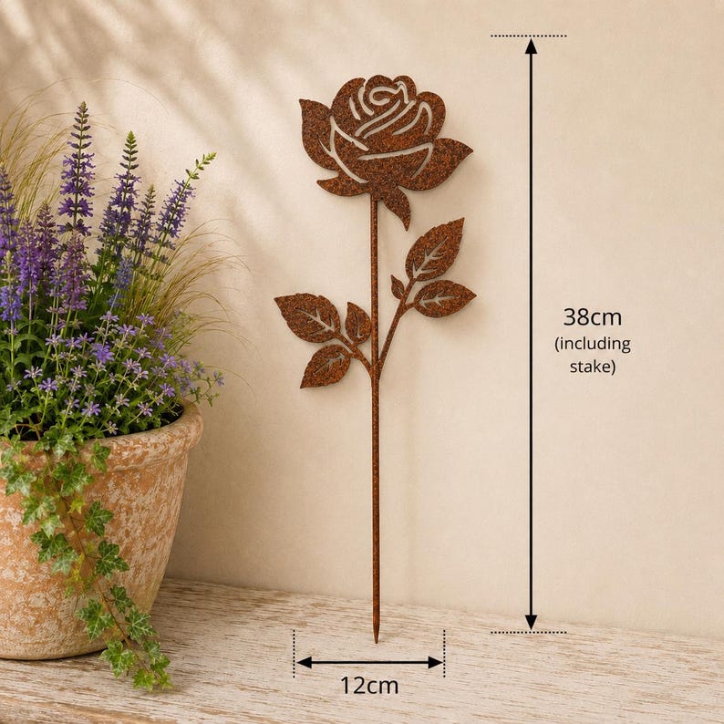 May include: A rust-colored metal rose garden stake featuring a cutout flower design with leaves on a thin stem. The total height is 38 cm including the stake, and the width is 12 cm. The metal has a textured, weathered brown finish throughout.