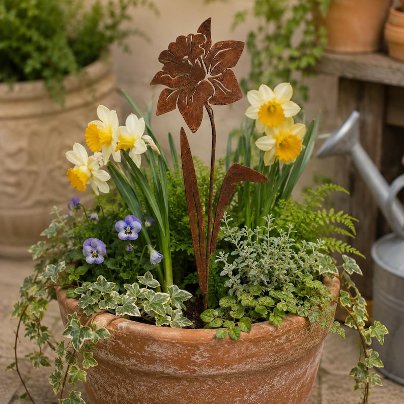 May include: A rust-colored metal daffodil garden stake stands in a terracotta pot filled with live yellow and white daffodils, purple pansies, and green ivy. The metal flower features a cutout design and a long stem, contrasting with the organic plants.
