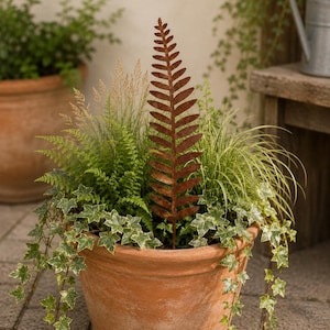 May include: A tall, slender, rust-colored metal fern leaf garden stake stands upright in a terracotta pot filled with green ferns, variegated ivy, and ornamental grasses. The metal fern features a detailed, symmetrical frond design.