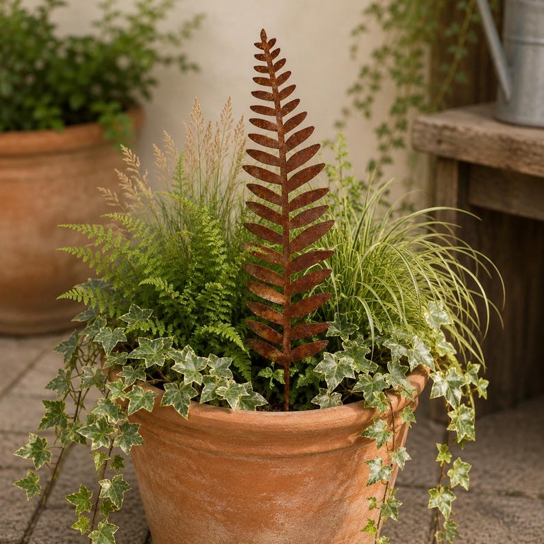 May include: A tall, slender, rust-colored metal fern leaf garden stake stands upright in a terracotta pot filled with green ferns, variegated ivy, and ornamental grasses. The metal fern features a detailed, symmetrical frond design.