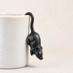 May include: A black cast iron mouse figurine designed to hook onto the rim of a mug. The mouse is depicted in a downward-facing position with its long tail curved upward, featuring a textured surface and detailed ears, eyes, and snout.