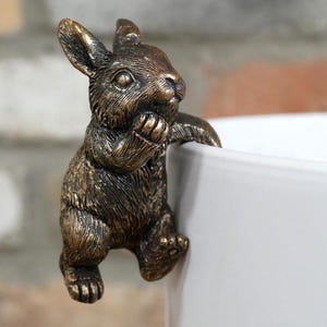 May include: A bronze-effect bunny rabbit ornament clinging to the edge of a white ceramic container. The rabbit has detailed fur texture and is posed with its paws resting on the rim. The background features a brick wall.